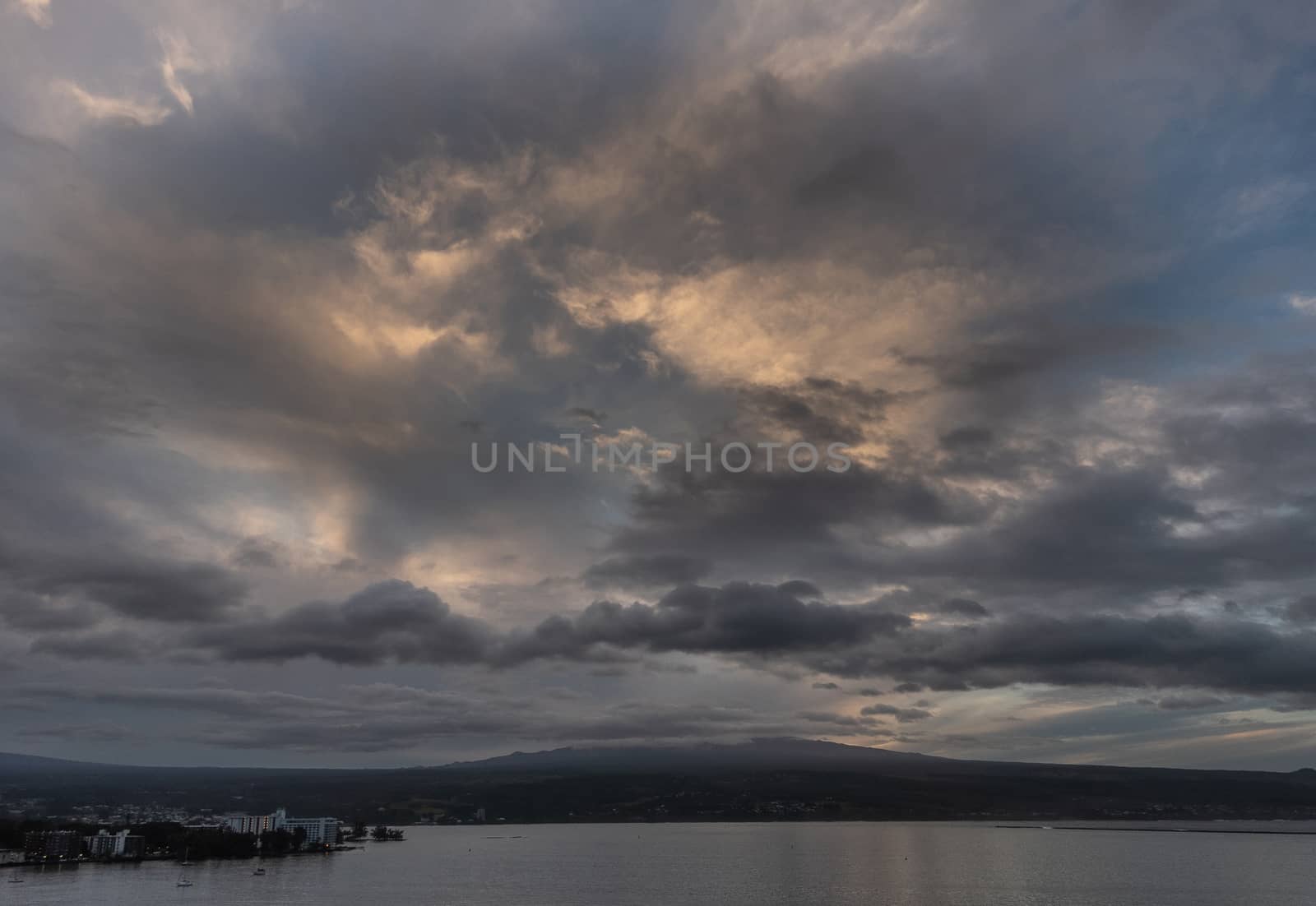 Hilo, Hawaii, USA. - January 14, 2020: Spectacular dark evening cloudscape with yellow sunlight patches over town and volcano on horizon.