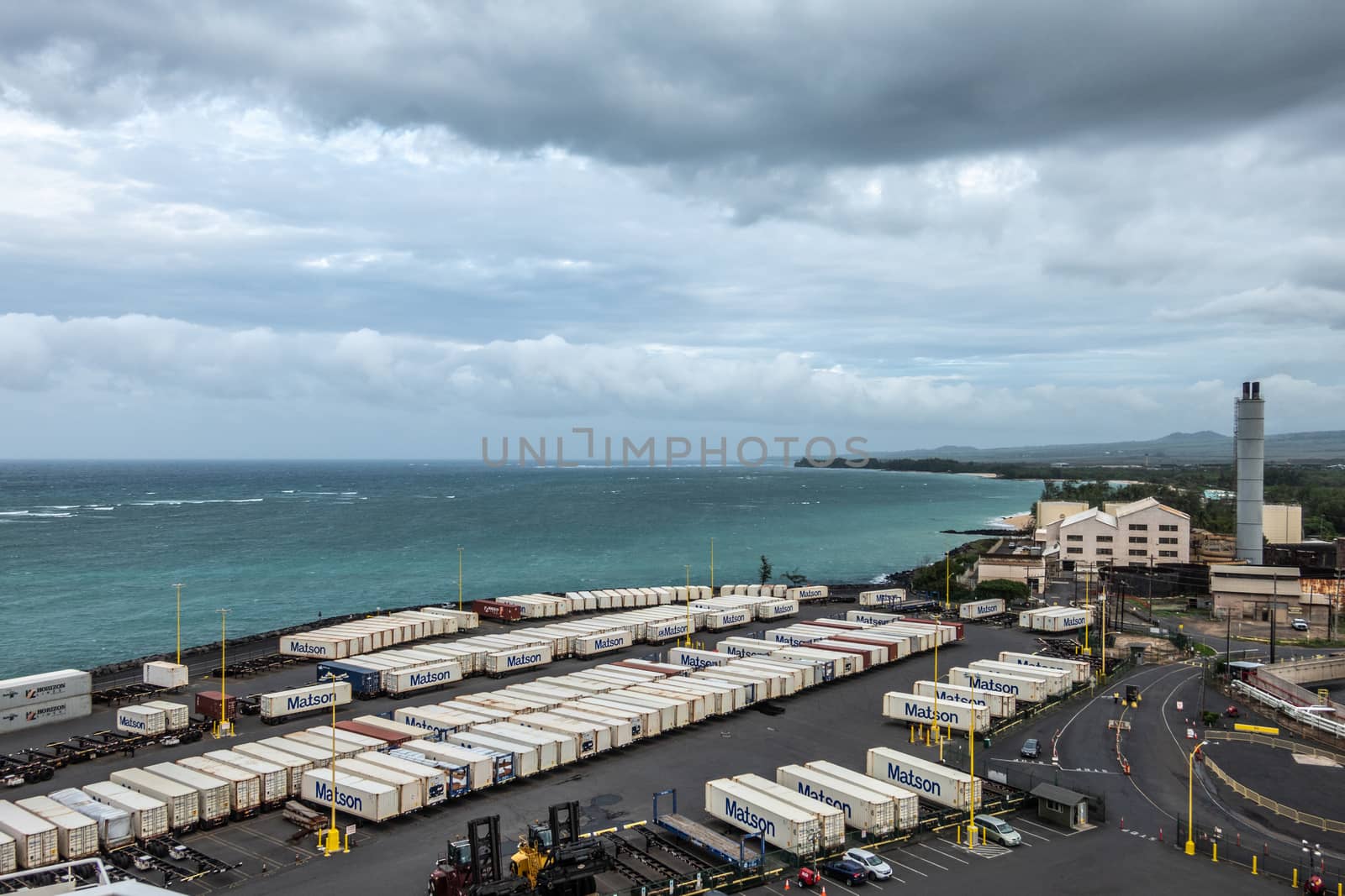 Kahului, Maui,, Hawaii, USA. - January 13, 2020: Matson shipping container yard full of white boxes on quay bordering azure ocean under gray rainy cloudscape. Old sugarcane processing plant.