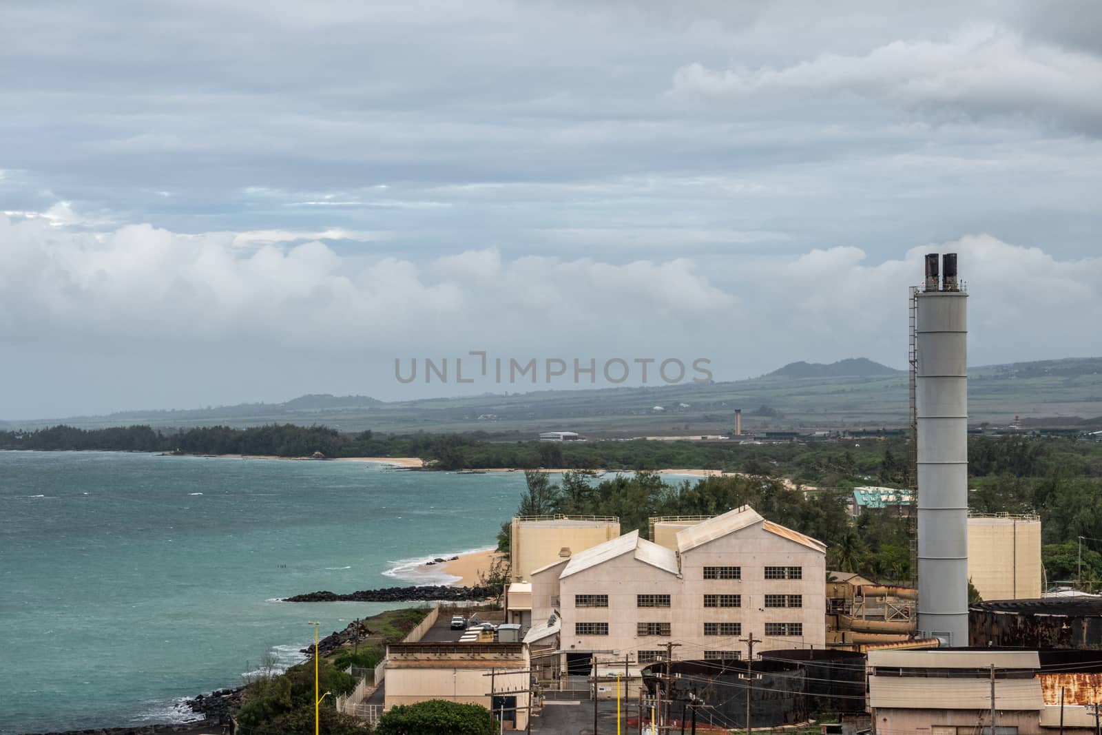 Kahului, Maui,, Hawaii, USA. - January 13, 2020: Old defunt sugarcane processing plant in harbor area bordering the azure ocean, under cloudscape with green hills and forest in back.