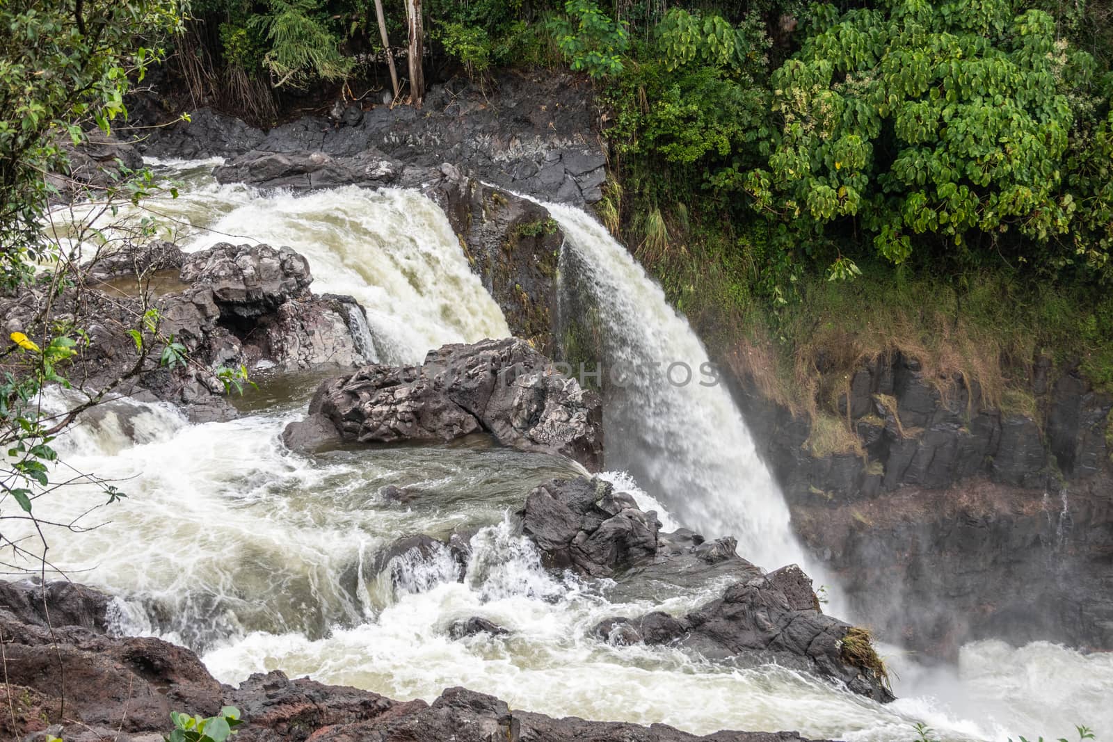 Hilo, Hawaii, USA. - January 14, 2020: Closeup of White water falling over lava rocks on edge of Rainbow Falls. Green vegetation.