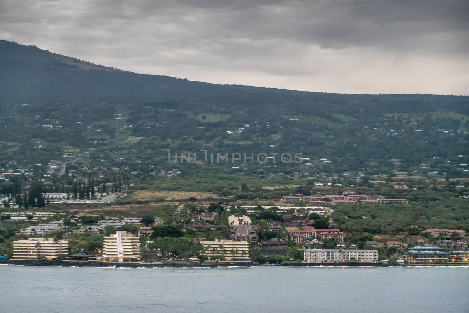 Kona, Hawaii, USA. - January 15, 2020: Resort hotel buildings along coastline set in green trees with forested mountain in back, dotted with red or white house roofs. Gray ocean in front.