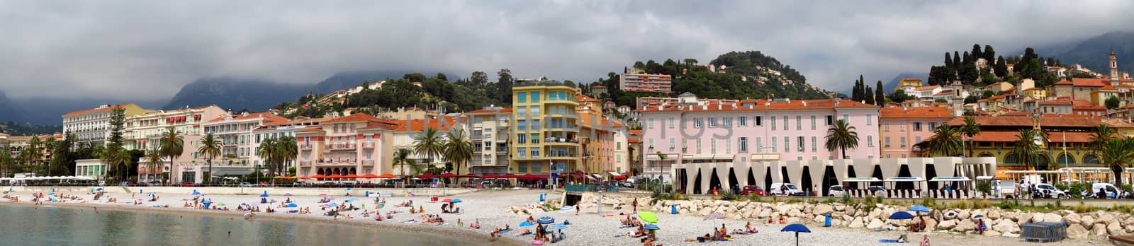 Menton, France - June 30, 2018: Menton beach and a panoramic view of the city. Unidentified people rest on the shore. Menton is a small seaside town on the French Riviera.