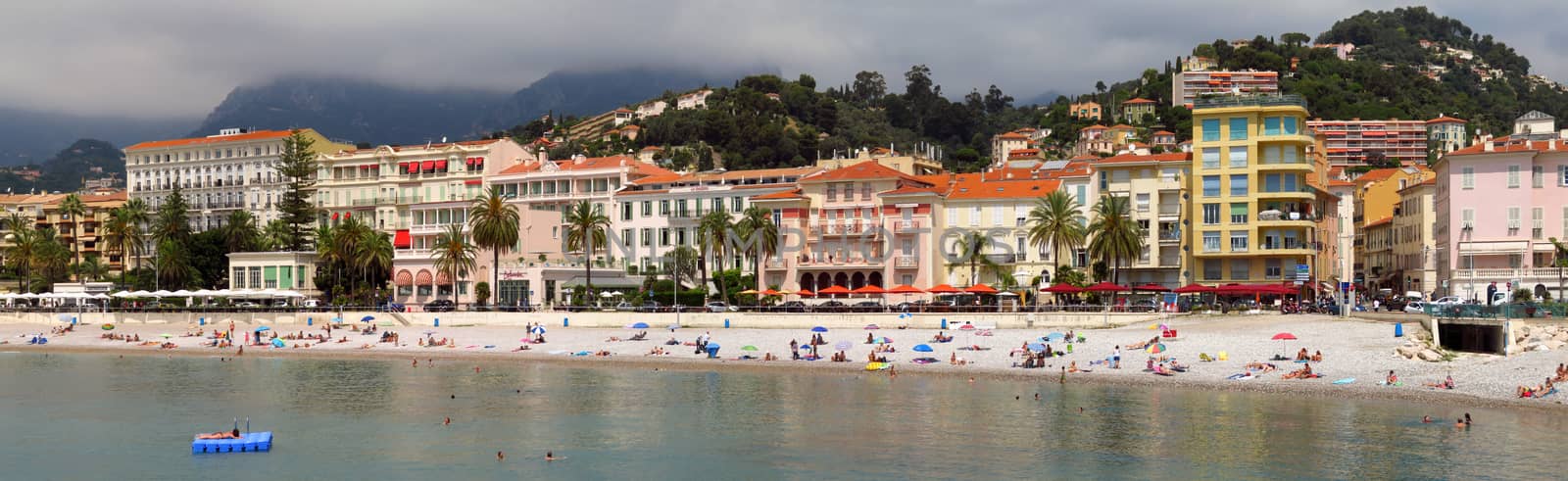 Menton, France - June 30, 2018: Menton beach and a panoramic view of the city. Unidentified people rest on the shore. Menton is a small seaside town on the French Riviera.