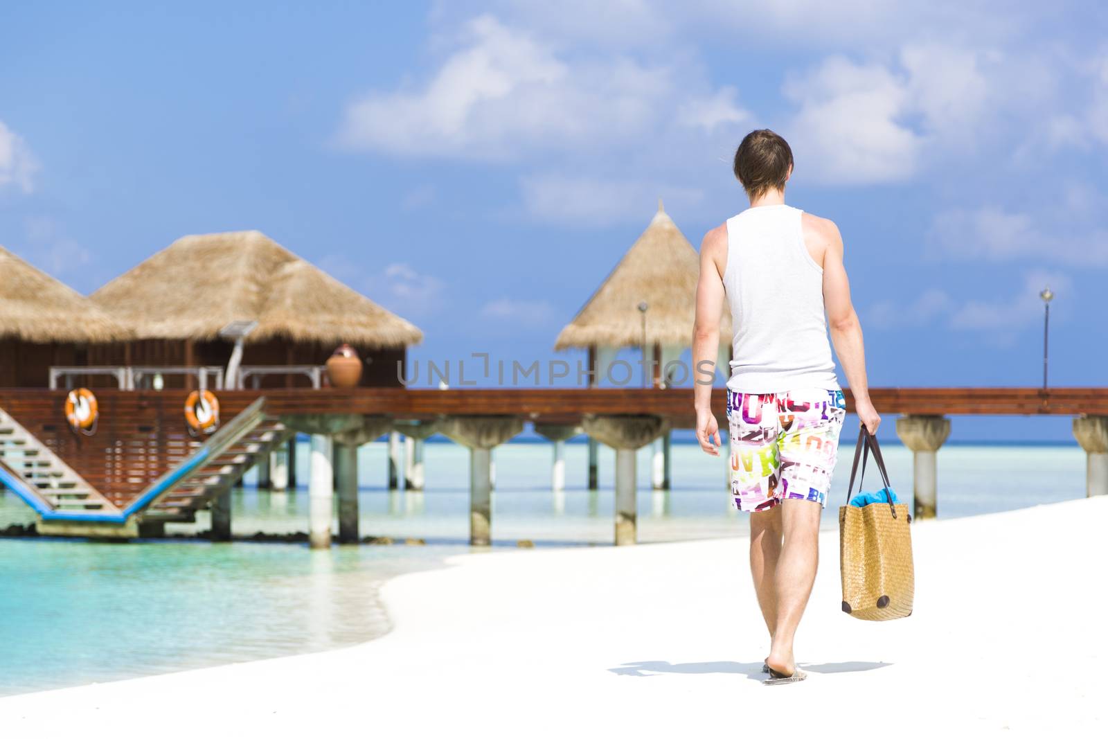 Man walking by the shore in the beach going away to a jetty with bungalows. He is wearing a colorful bathing suit while carrying a beach bag with towels. At background there are bungalows out of focus.