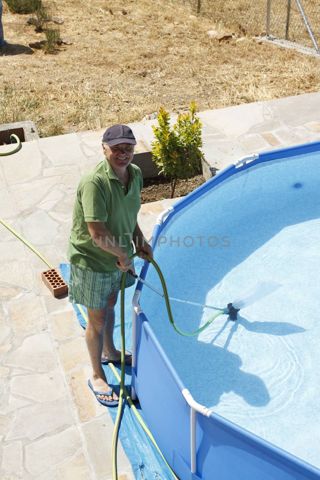 A senior man smiles while cleaning up the swimming pool at his garden.