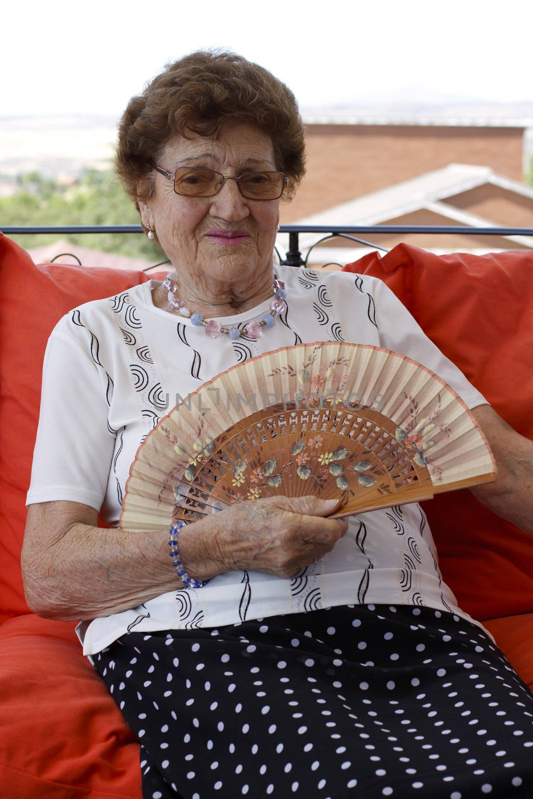 Old woman sitting in the patio in red cushion with a hand fan.