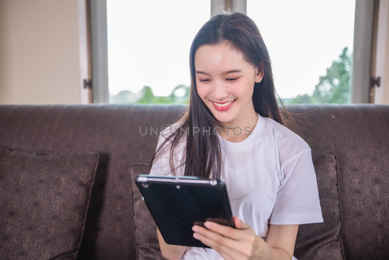 Asian young woman happy cheerful cute beautiful is playing digital pad tablet computer and sitting on the sofa at home, smiling lady using app reading electronic book relax in living room