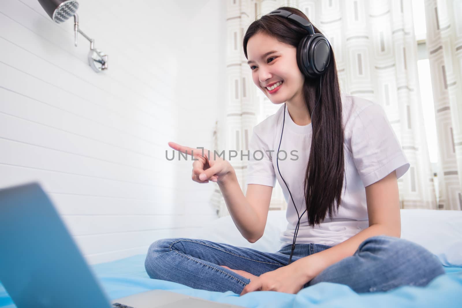 Asian happy young woman wear headphones and listening music song and watching website for shopping online from laptop  on bed at home