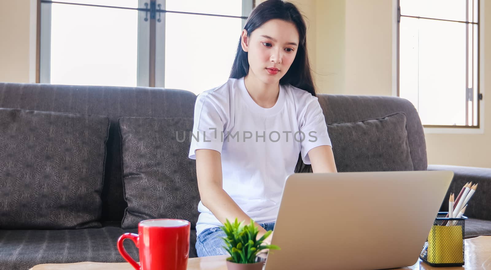 Asian woman happy cheerful cute beautiful is using a notebook computer and sitting on sofa for working from home. She is quarantined at home during the coronavirus epidemic.