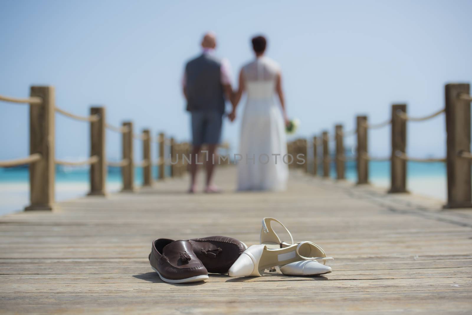 Married couple on wedding day walking romantically outside on wooden pier with pair of shoes in foreground