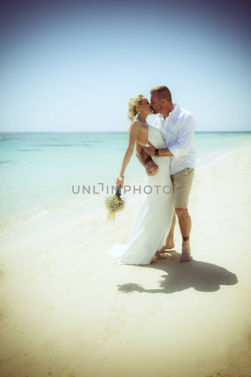 Beautiful married couple kissing at a tropical beach paradise on wedding day in white gown dress with ocean view vintage style photo