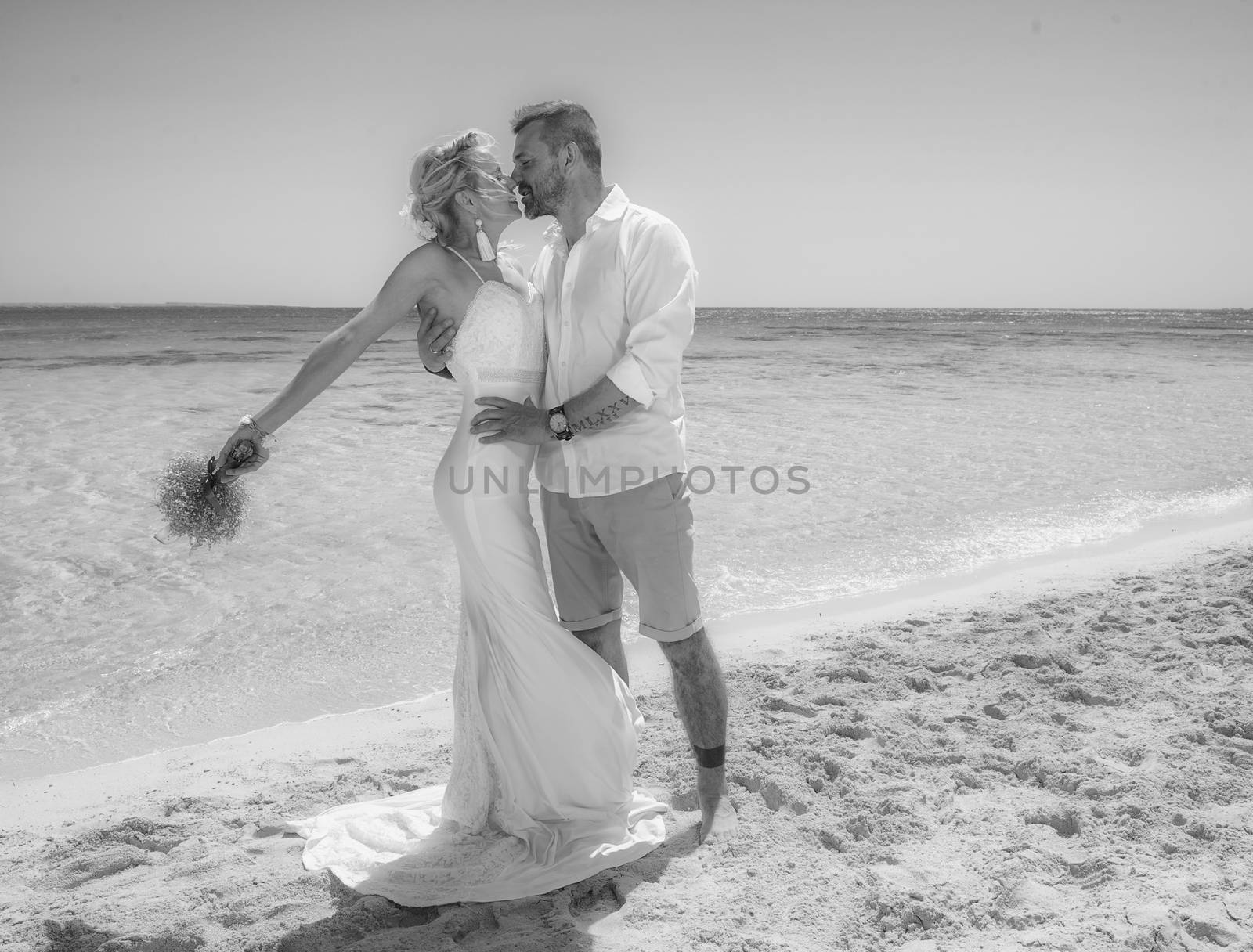 Beautiful married couple kissing at a tropical beach paradise on wedding day in gown dress with ocean view black and white photo