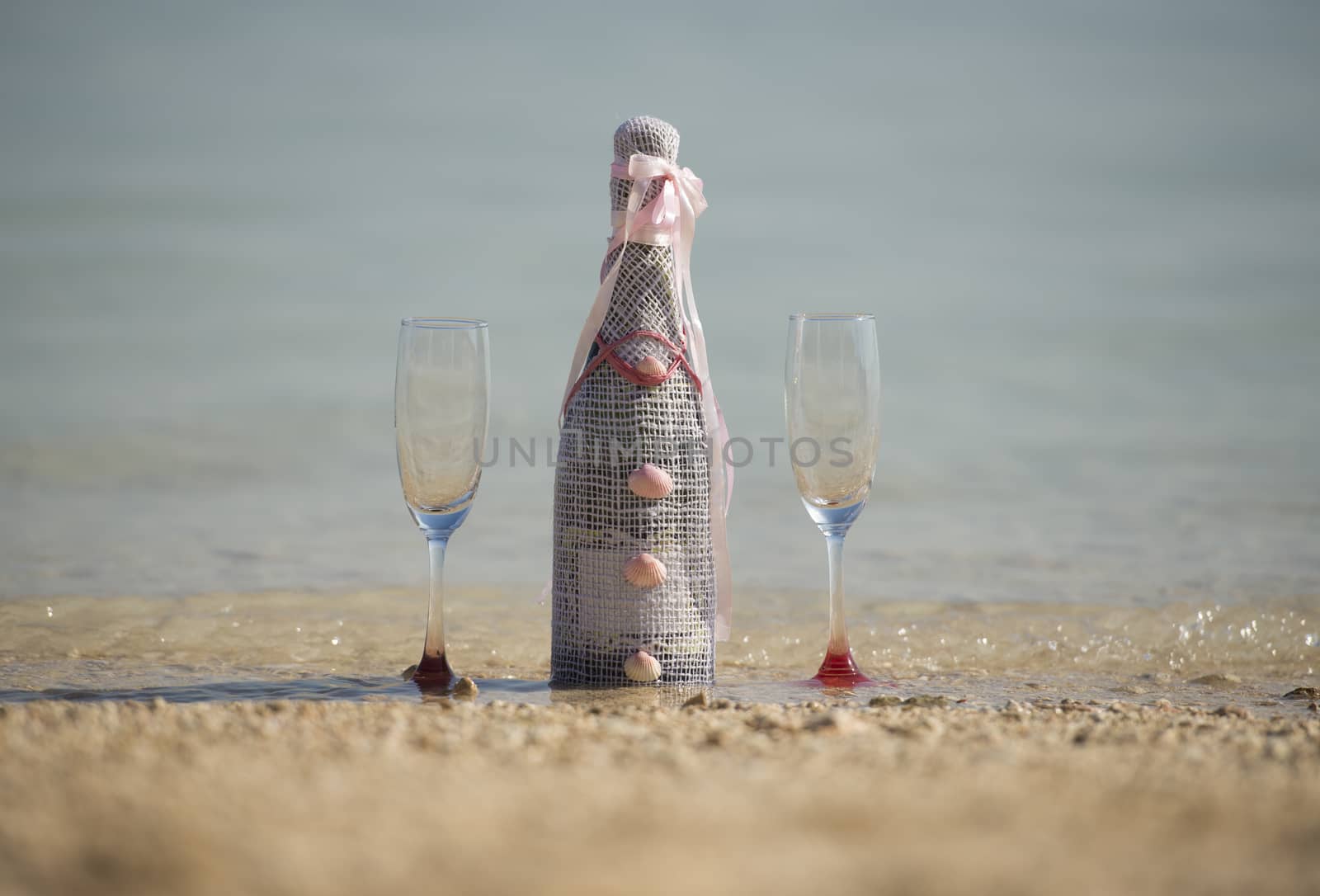 Decorated bottle of champagne sparkling wine and two glasses on tropical sandy beach in the water romantic concept
