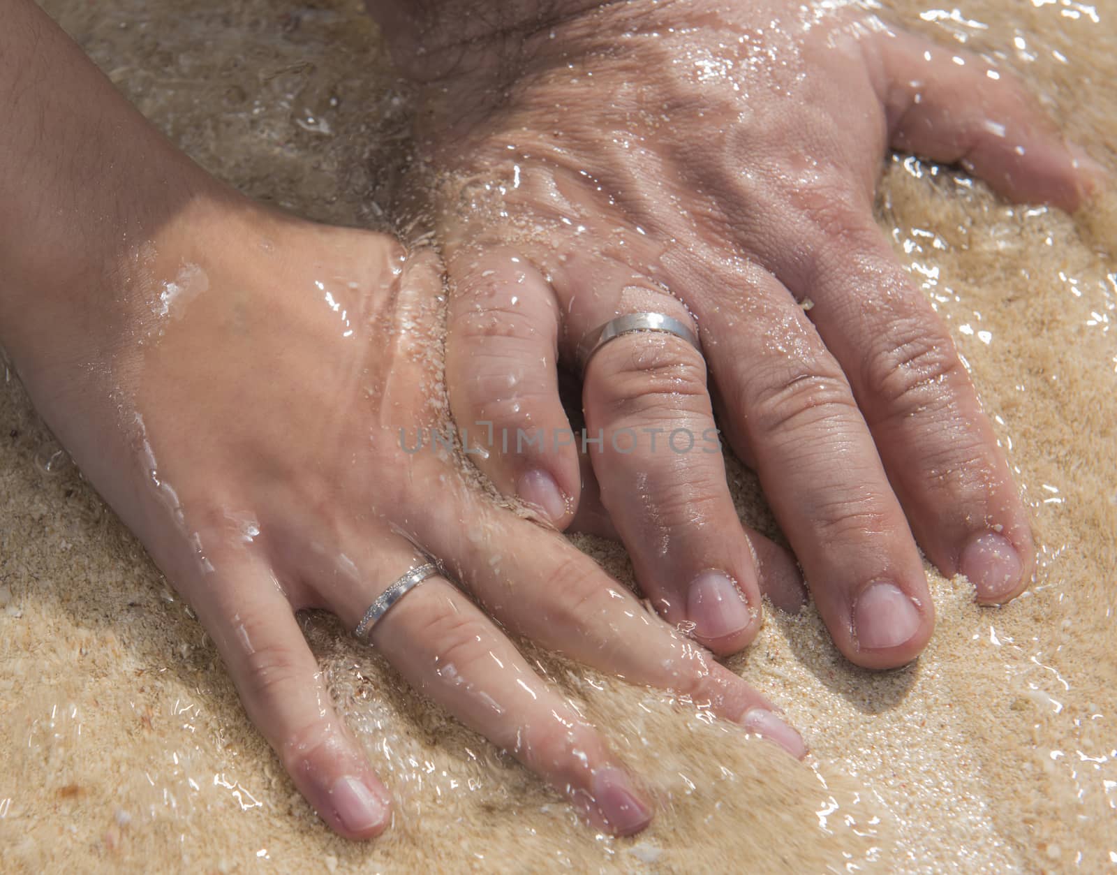 Hands of bride and groom on wedding day showing rings at tropical sandy beach in water romantic concept