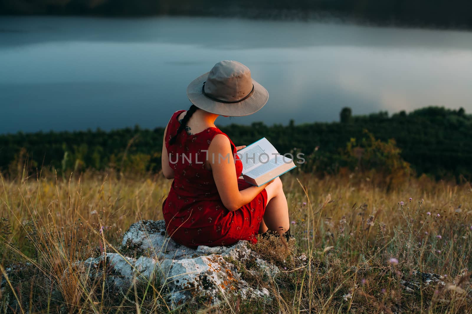 Girl sitting on a stone in the mountains reads a book against the backdrop of a large river. Portrait at sunset. The girl in the hat. Holidays at the hotel