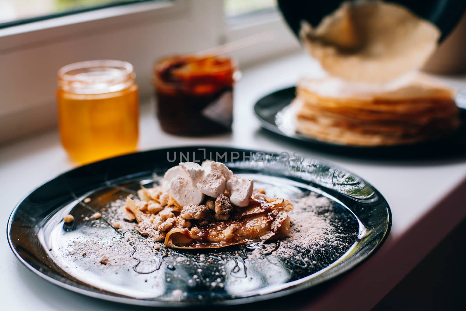 Pancakes with caramel nuts and marshmallows on the kitchen windowsill. In the background are a lot of pancakes, caramel and honey in a jar.