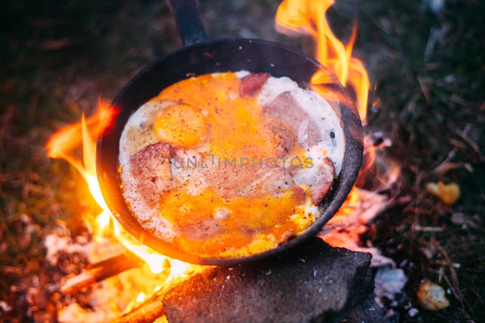 Fried eggs with bacon in a pan in the forest. Food at the camp. Scrambled eggs with bacon on fire. Lots of seasonings on fried eggs. Picnic