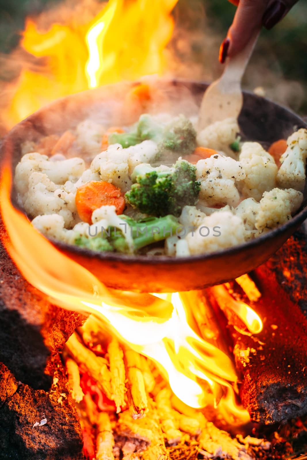 Cauliflower, broccoli and carrot in a pan. Cooking on an open fire. Outdoor food. Grilled vegetables. Food on a camping trip. Hand with a spatula