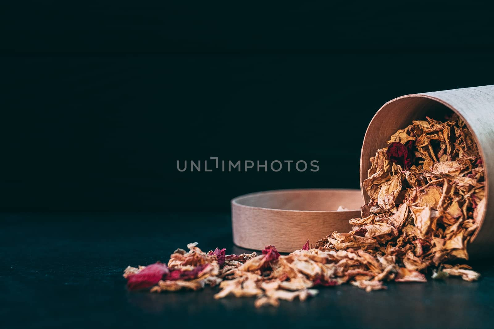 Scattered tea rose petals with a wooden jar on a dark background by Opikanets