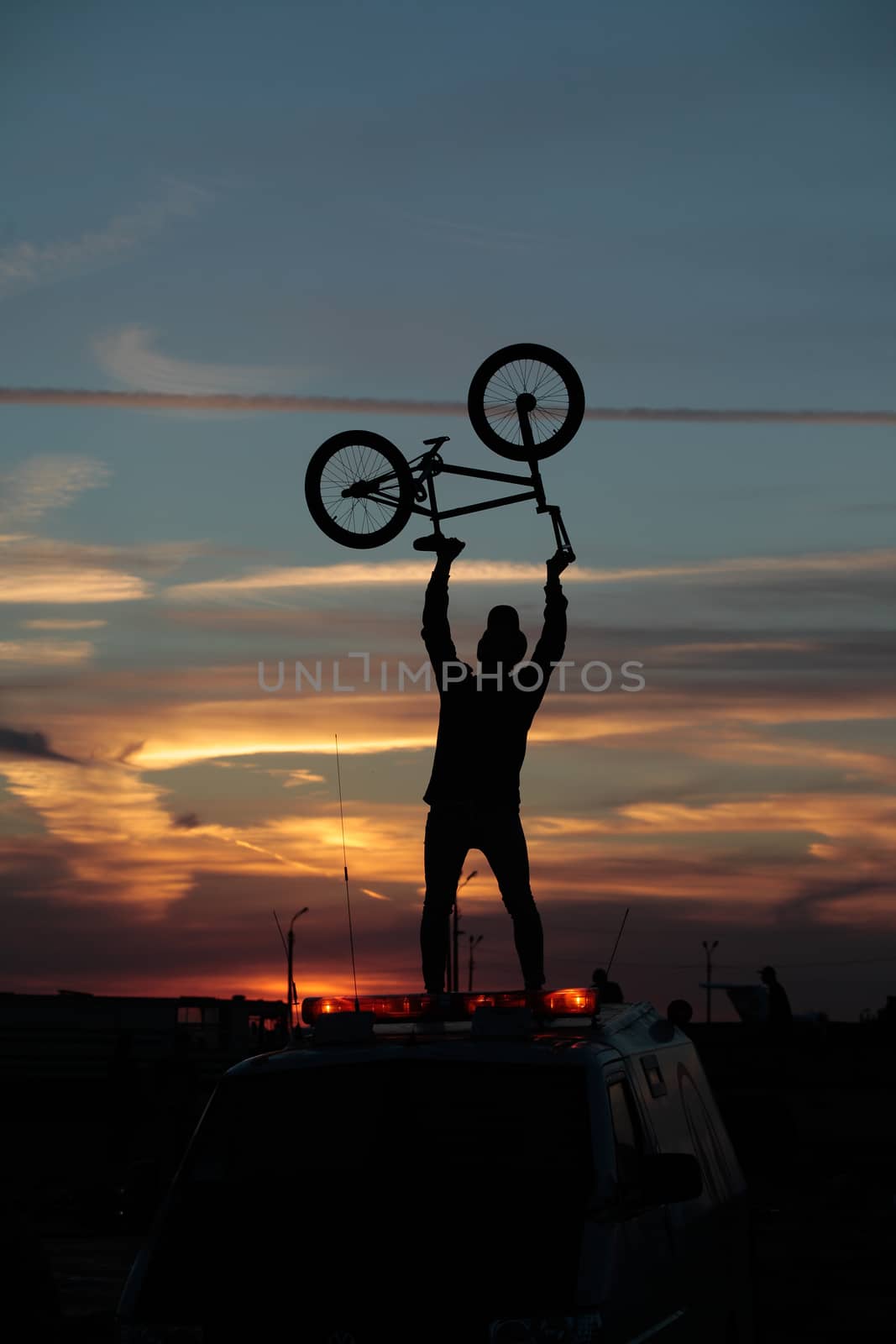 Silhouette of a cyclist holding his bike over his head on a sunset background, standing by a car