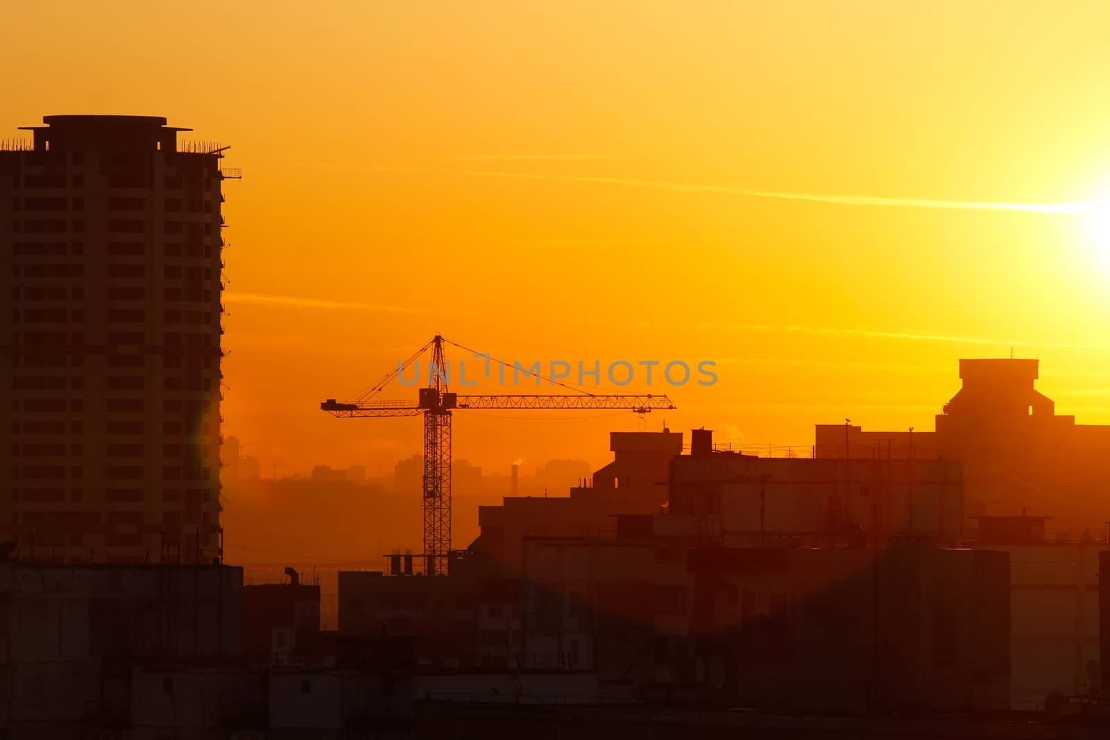 Orange sunset on the background of the city. Silhouette of a construction crane. Photo with a glare from the sun. Panorama of the city.