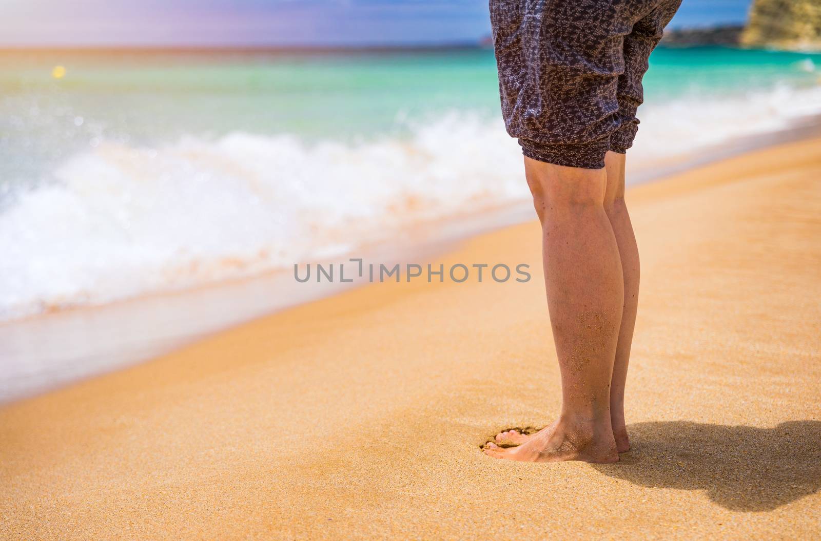 Closeup on leg of young woman standing on sea shore. Woman legs and feet walking on the sand of the beach with the sea water in the background. Woman standing on the beach with wave sea.