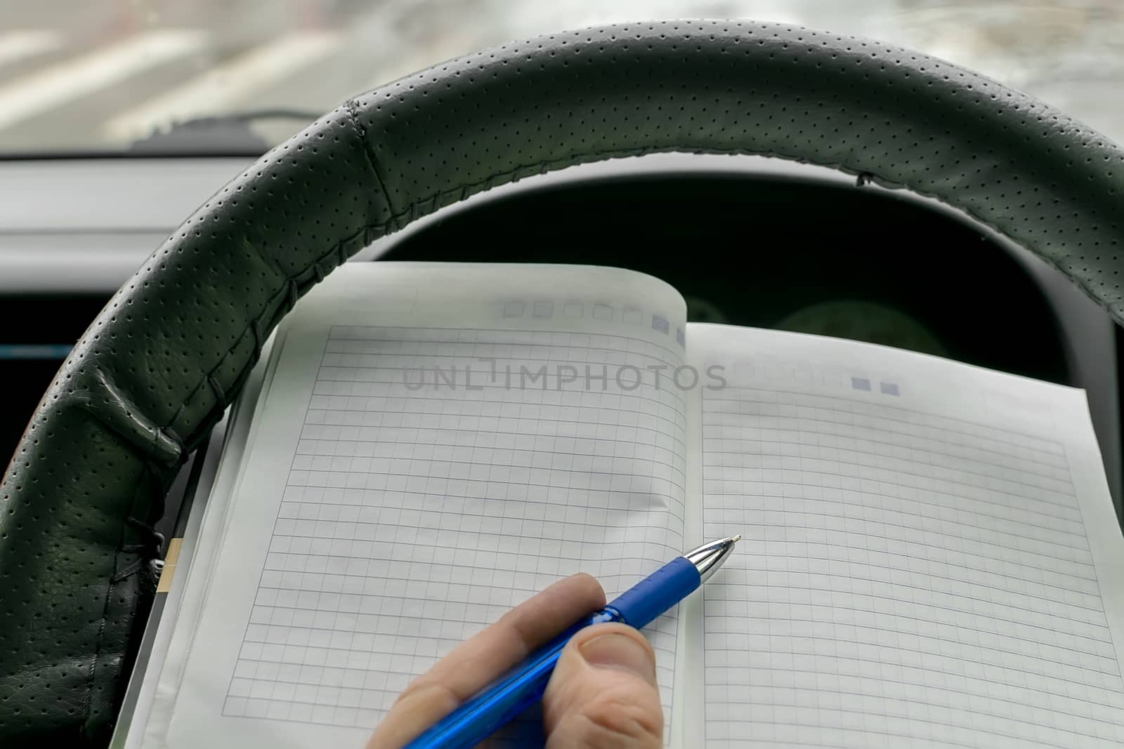 the hand of the car driver behind the wheel holds a paper notebook and makes notes