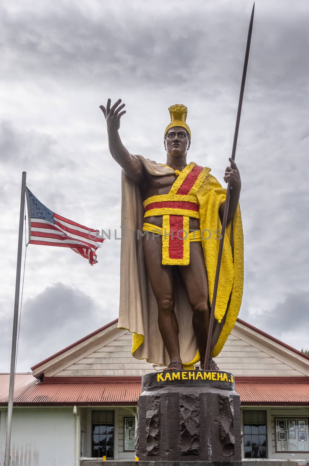 Kapaau, Hawaii, USA. - January 15, 2020: Closeup of statue of King Kamehameha, decorated with yellow and red dress, golden crown and long spear under gray cloudscape with USA flag behind.