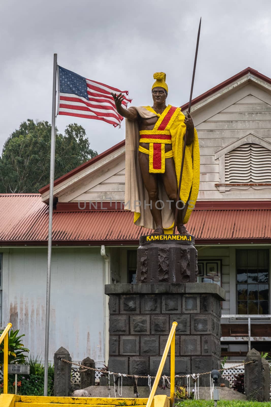 Kapaau, Hawaii, USA. - January 15, 2020: Statue of King Kamehameha, decorated with yellow and red dress, golden crown and long spear under gray cloudscape with USA flag behind.