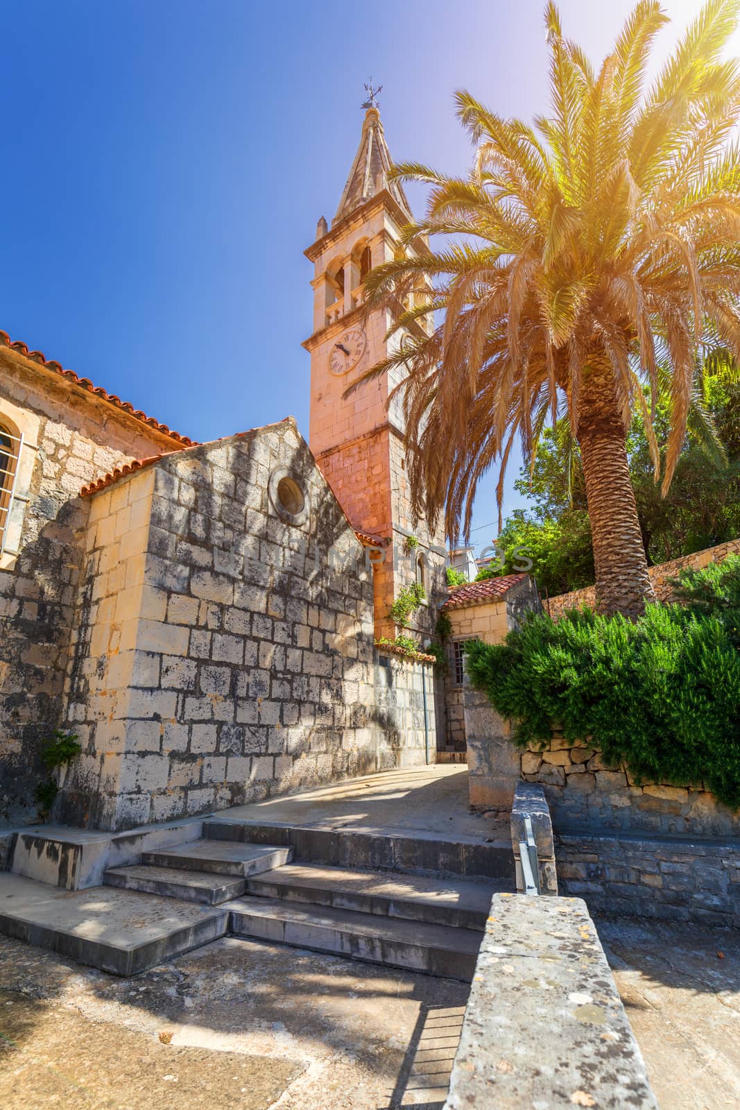 Church building and palm tree against sunny blue sky in Splitska village on Brac island, Croatia. 