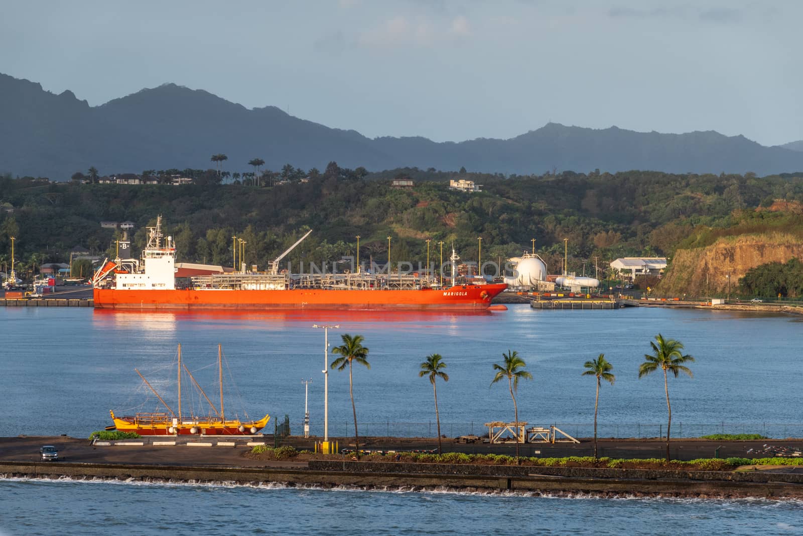 Nawiliwili, Kauai, Hawaii, USA. - January 16, 2020: Early morning light on red Marigola LPG tanker in port under light blue sky. Green belt on hill. Blue ocean water and pier in front with historic sail boat.