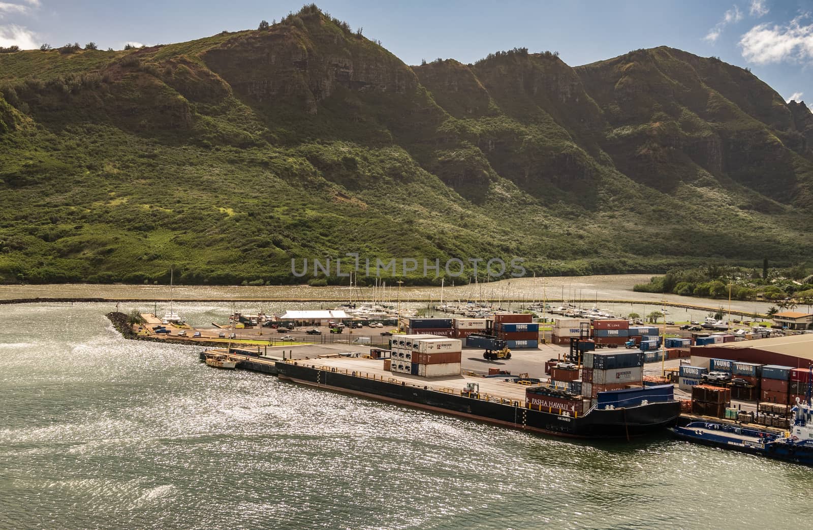 Nawiliwili, Kauai, Hawaii, USA. - January 17, 2020: Almost empty Haaheo shipping container barge docked in port. Green mountain with brown-black cliffs as backdrop under light blue cloudscape.