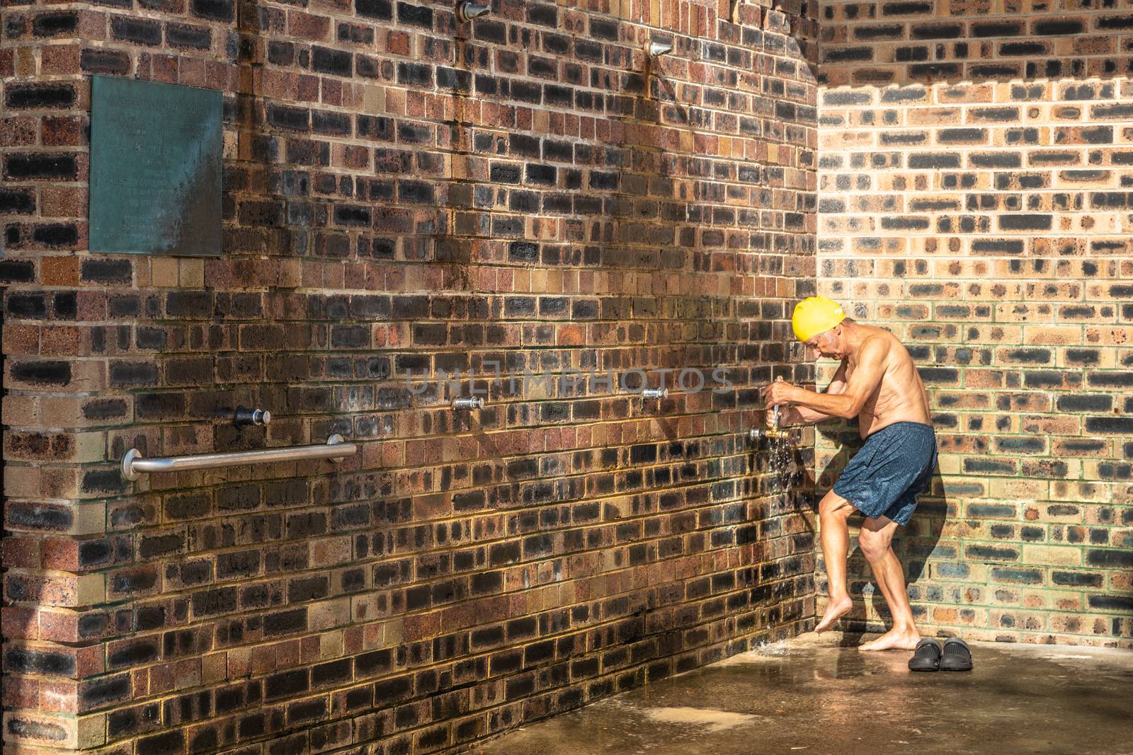 Sydney, Australia - February 11, 2019: Older man with yellow cap rinses feet with water in brown-stone shower facility at Bronte Beach.