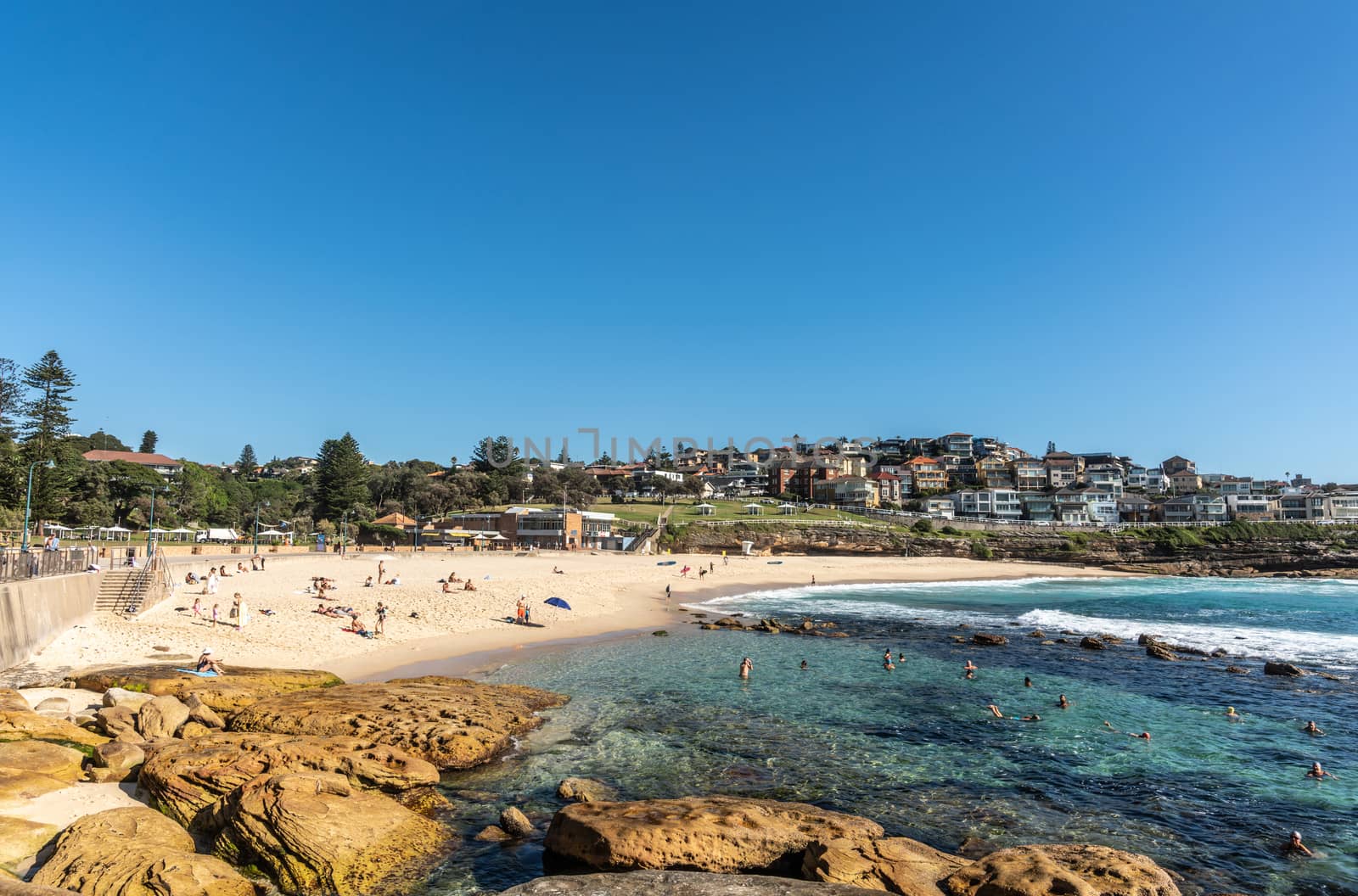 Sydney, Australia - February 11, 2019: Bronte Beach, sea water, yellow sand, green park under blue sky, seen from South cliff. Band with housing on horizon. Brown rocks in foreground. People active.