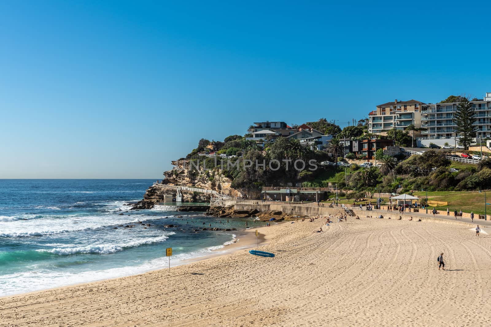 Sydney, Australia - February 11, 2019: Bronte Beach, sea water, yellow sand, green park under blue sky seen from North shore.. Band with housing on horizon. People active.