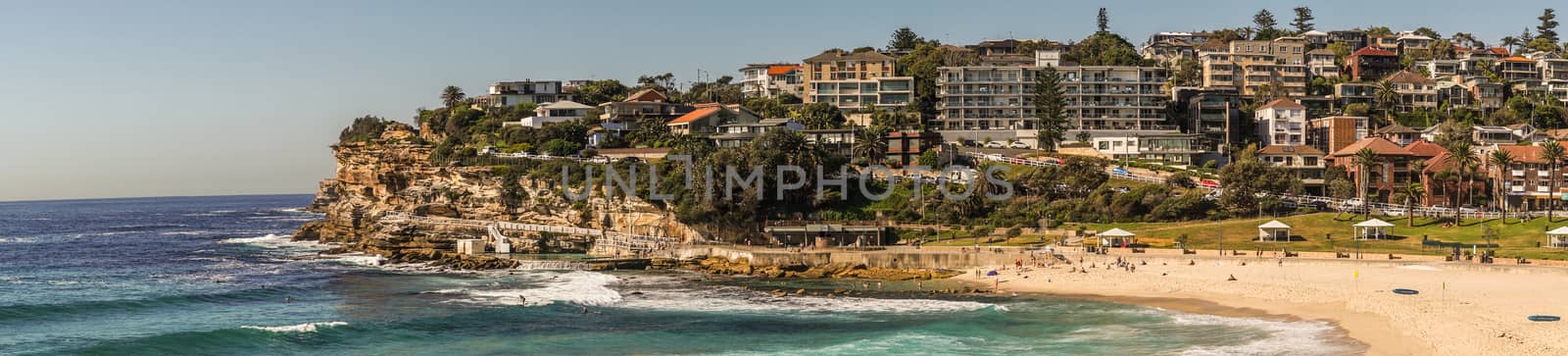 Sydney, Australia - February 11, 2019: Panorama shot of Bronte Beach, sea water, yellow sand, green park under blue sky, seen from North cliff. Band with housing on horizon. Brown rocks in foreground. People active.