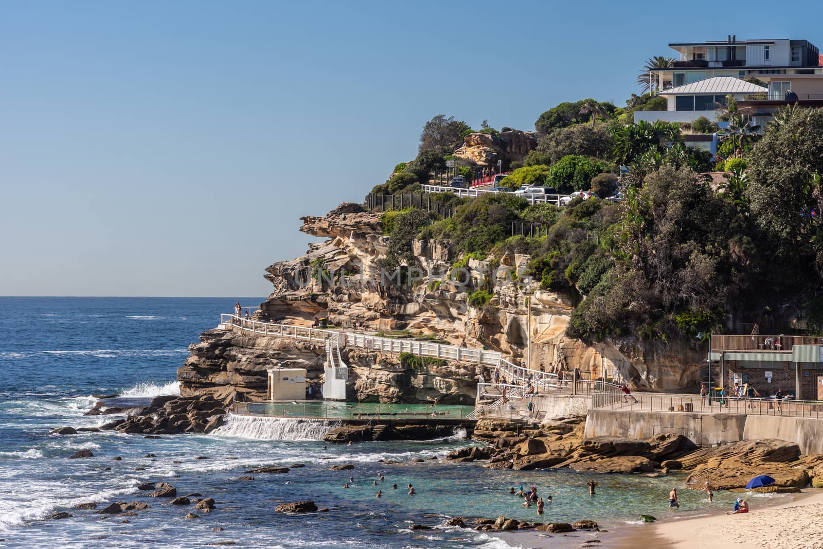 Sydney, Australia - February 11, 2019: Focus on the South Cliff at Bronte Beach with swimming pool in front. Peple in water and on sand. Housing in green vegetation on top of rocks.