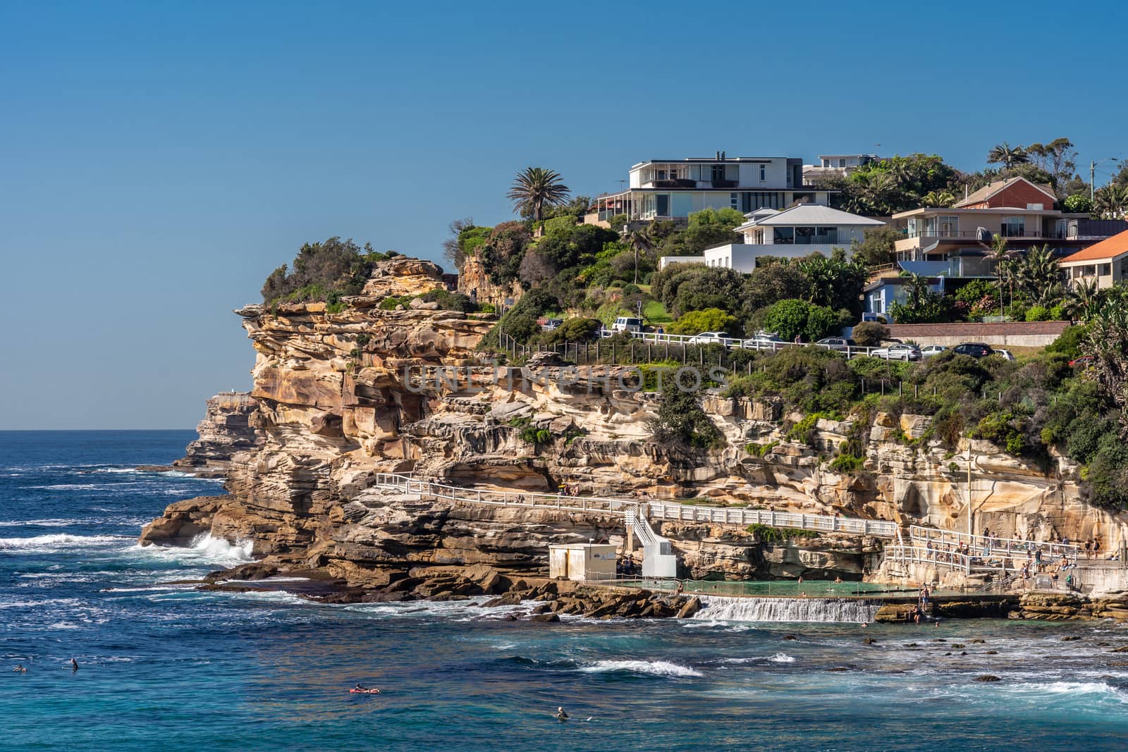 Sydney, Australia - February 11, 2019: Focus on the South Cliff at Bronte Beach with swimming pool in front. Peple in water. Housing in green vegetation on top of rocks.