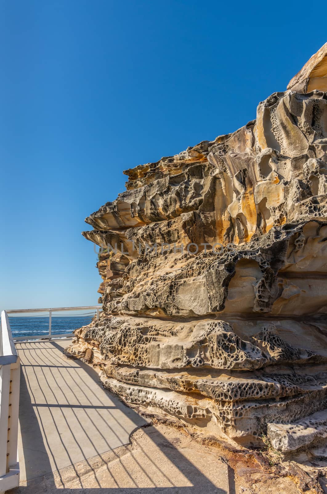 Sydney, Australia - February 11, 2019: Path ends at spectacular rock outcrop at Bronte Beach South cliffs, made by erosion.. Yellows and browns under blue sky. Sponge-like borders of shell like plates sticking out.