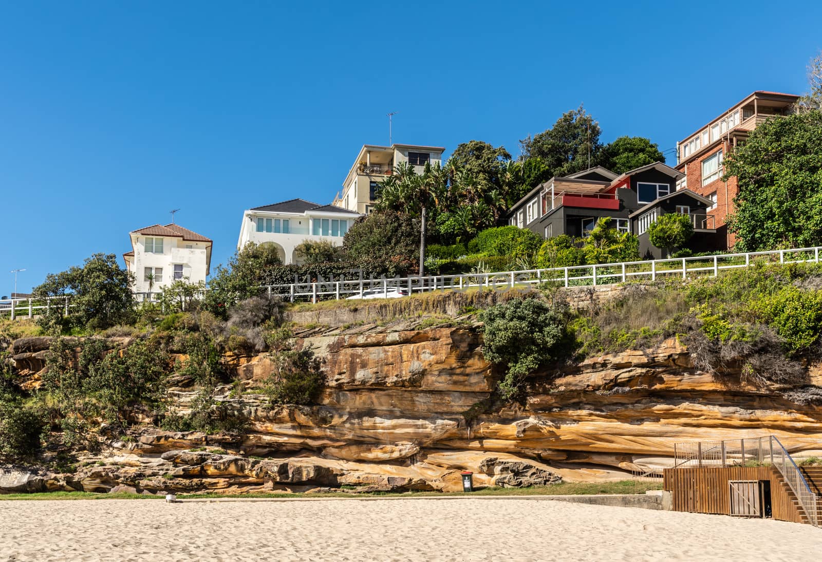 Sydney, Australia - February 11, 2019: The green overgrown cliffs, south side, with houses on top and sand in front of Tamarama beach under blue sky.