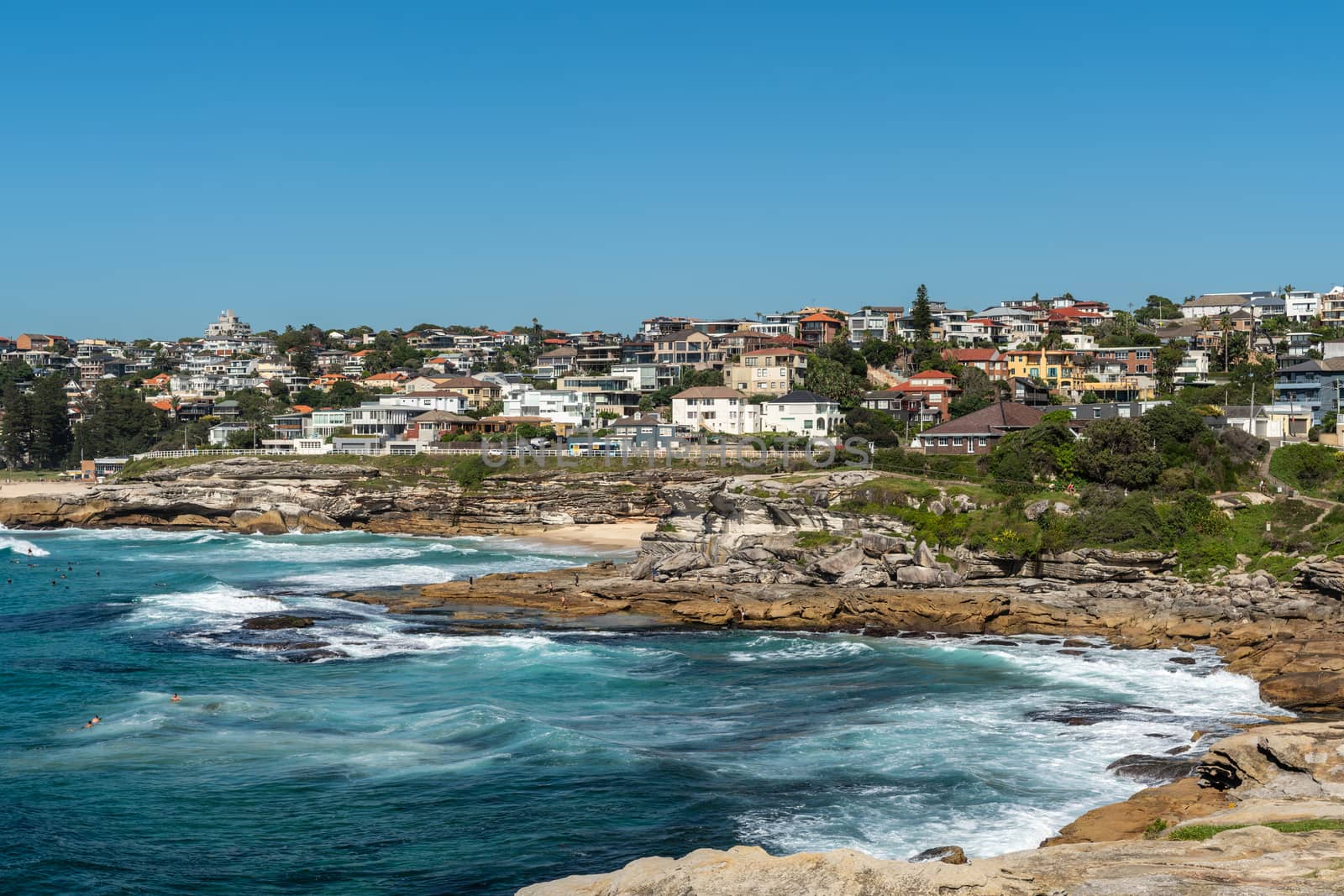Sydney, Australia - February 11, 2019: Wide shot of Bronte and Tamarama beaches with neighborhoods above and rocks to the north. Blue sea and blue sky. Waves crashing on rocks.