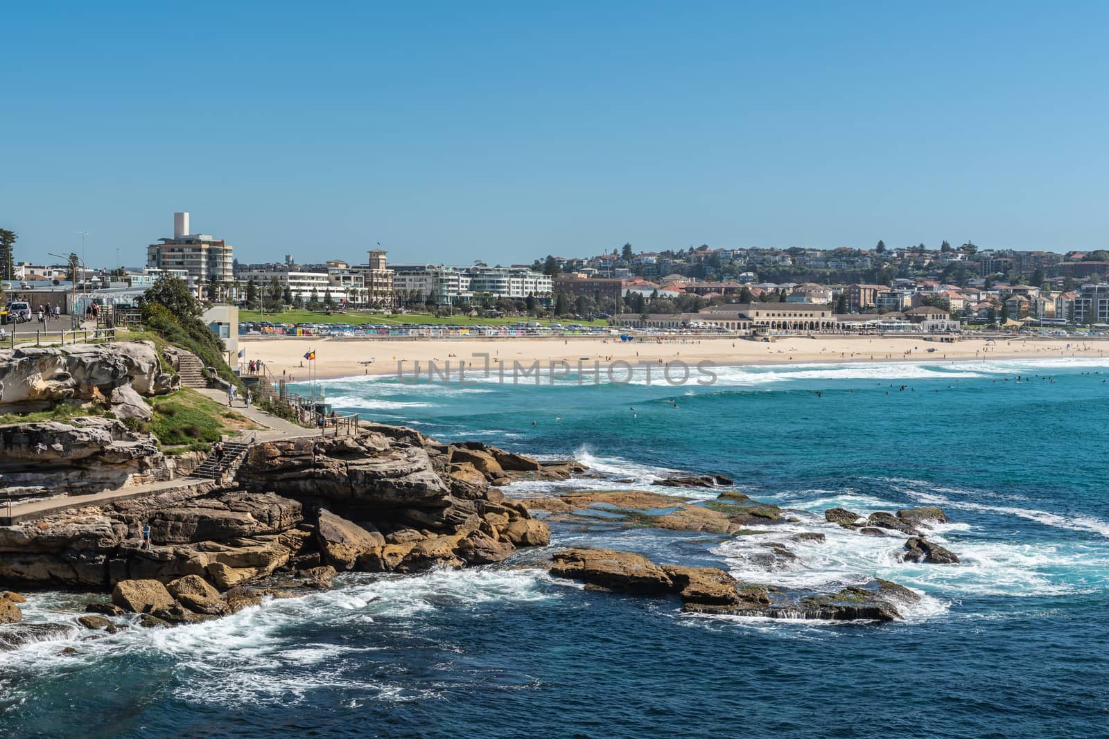Sydney, Australia - February 11, 2019:  Sandy Bondi beach with gray rocky Mackenzies Point in front. Horizon is band of buildings of Bondi. Blue water and blue sky.