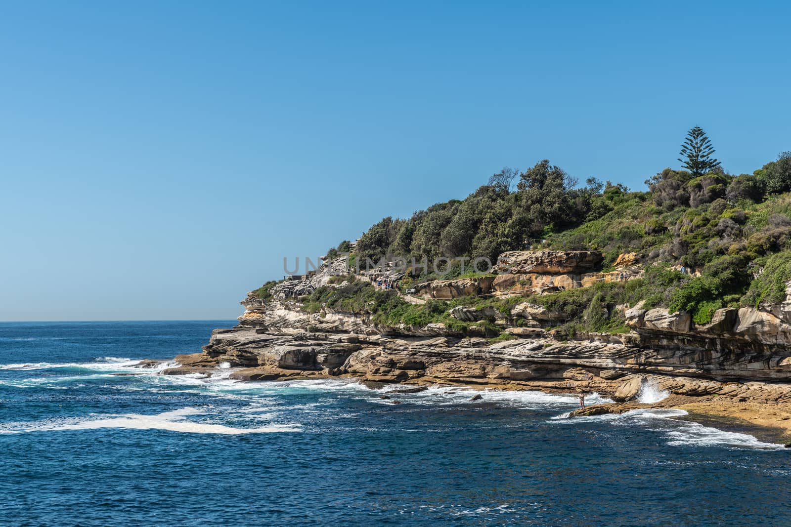 Sydney, Australia - February 11, 2019:  South side rocks and cliffs at Bondi beach. Green vegetation on top. People walking on path. Blue water and sky.