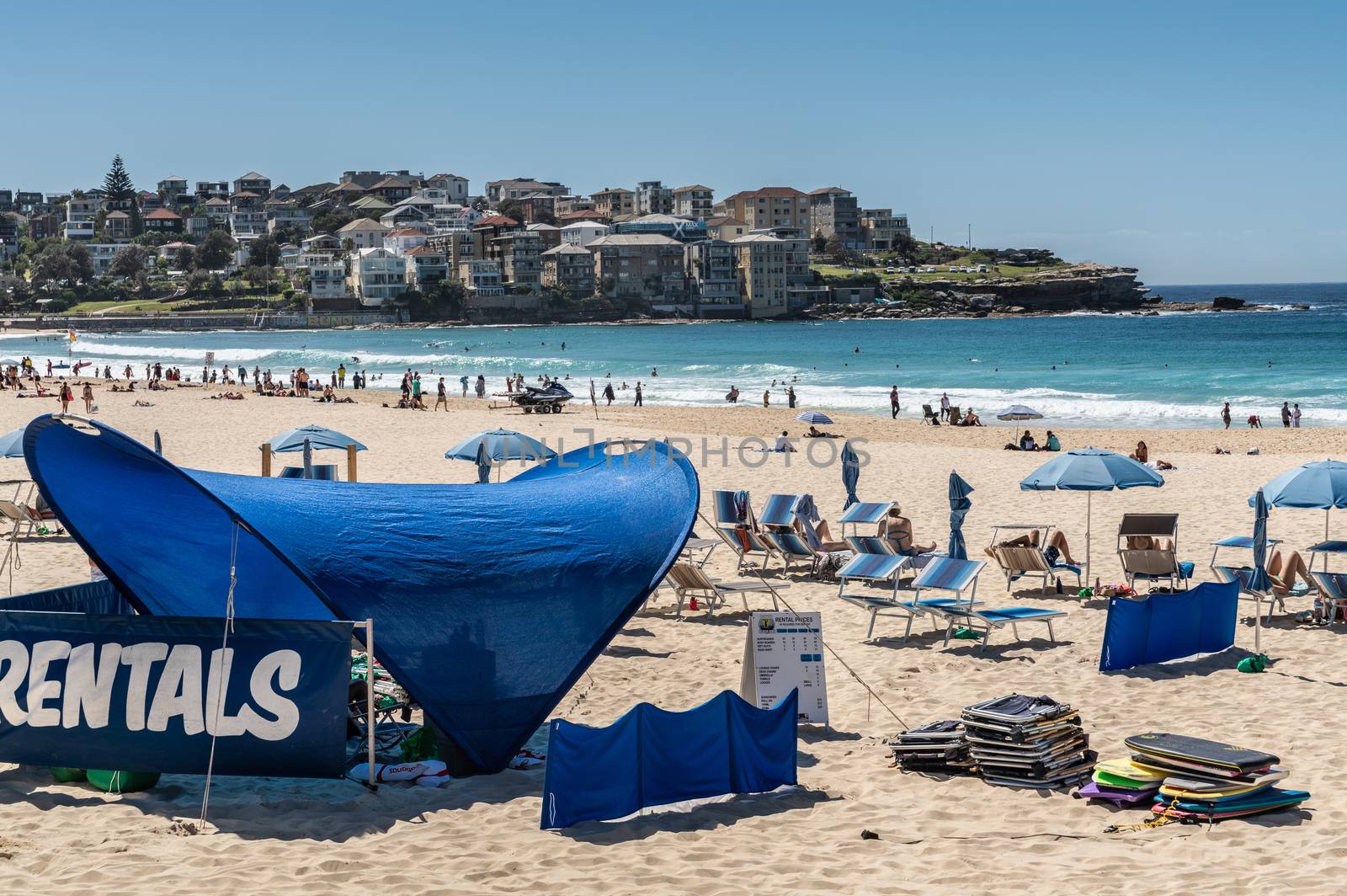 Sydney, Australia - February 11, 2019: Bondi Beach and North side cliffs with housing, under blue sky. Pale yellow sand with people, blue umbrellas and  beach beds and folding chairs in front, blue water.