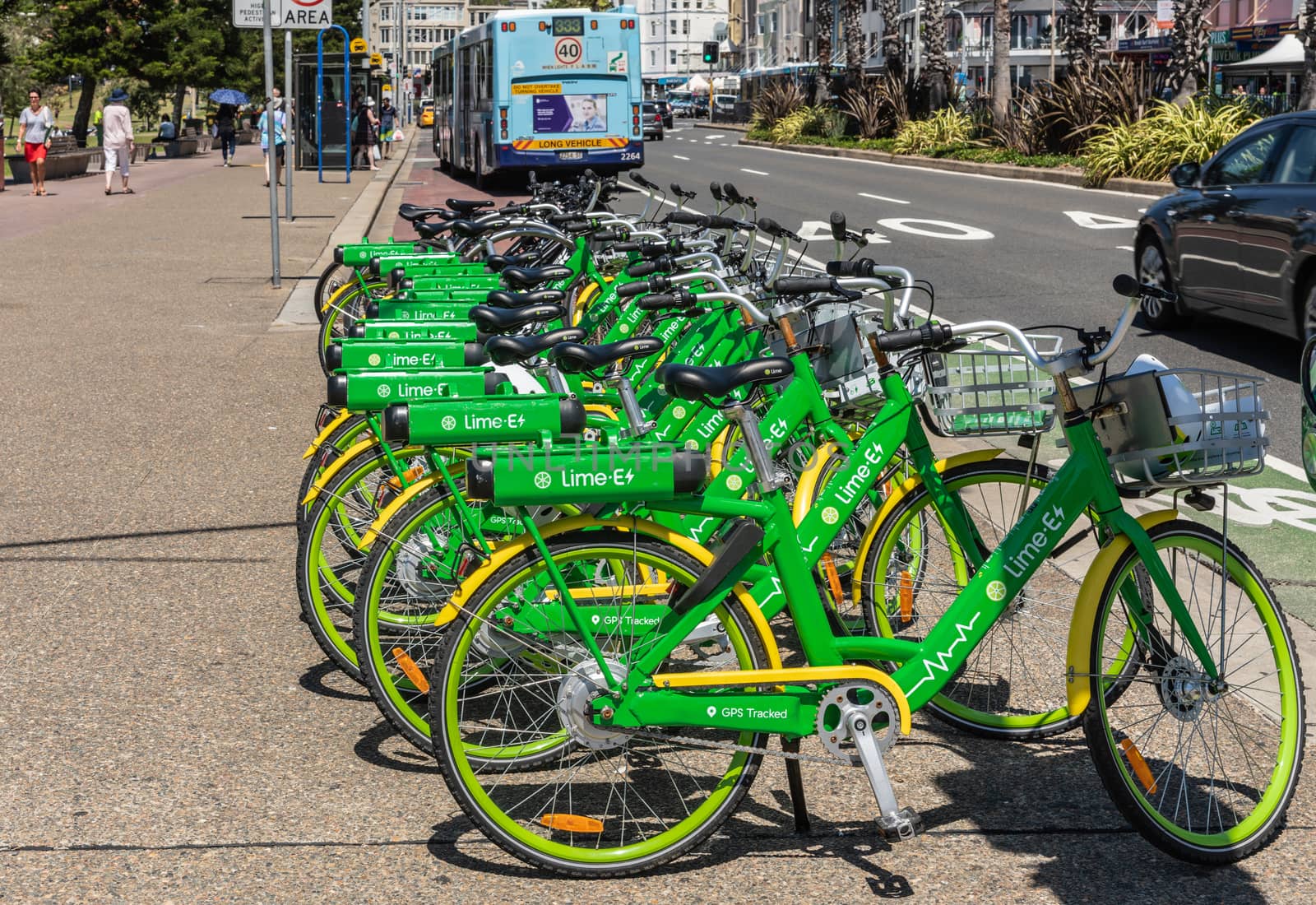 Sydney, Australia - February 11, 2019: Line of green and yellow Lime E-bicycles stationed on sidewalk of road bordering Bondi beach. People, bus and cars.