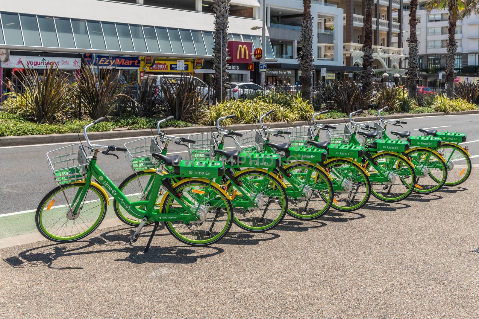 Sydney, Australia - February 11, 2019: Line of green and yellow Lime E-bicycles stationed on sidewalk of road bordering Bondi beach. People and cars.