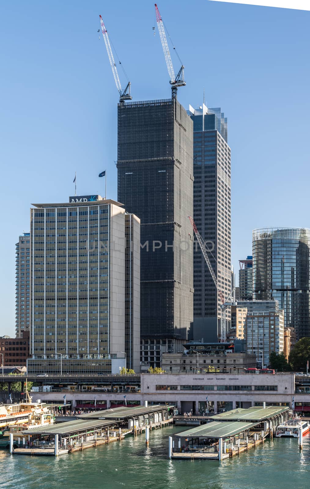 Sydney, Australia - February 12, 2019: Part of ferry terminal and Circular Quay Railway Station plus skyline in back. Highrises under construction with cranes. Evening shot with light blue sky.