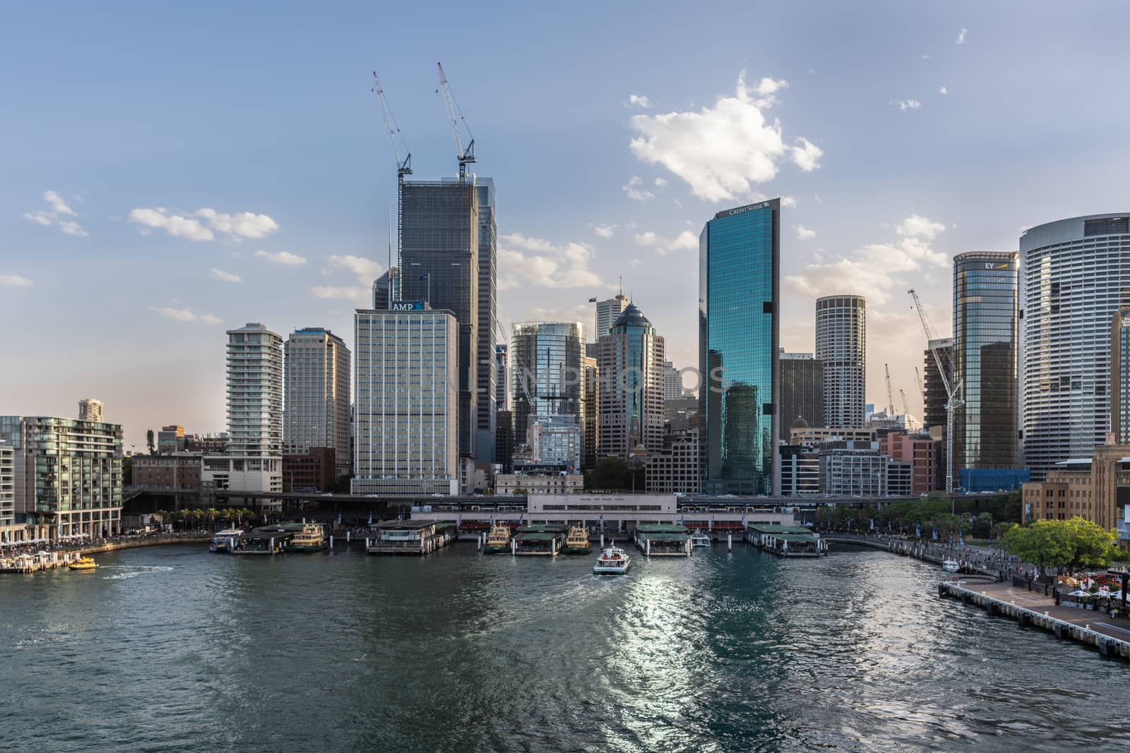 Sydney, Australia - February 12, 2019: Ferry terminal and Circular Quay Railway Station plus skyline in back. Highrises under construction with cranes. First Fleet Park green. Evening shot with light blue sky.