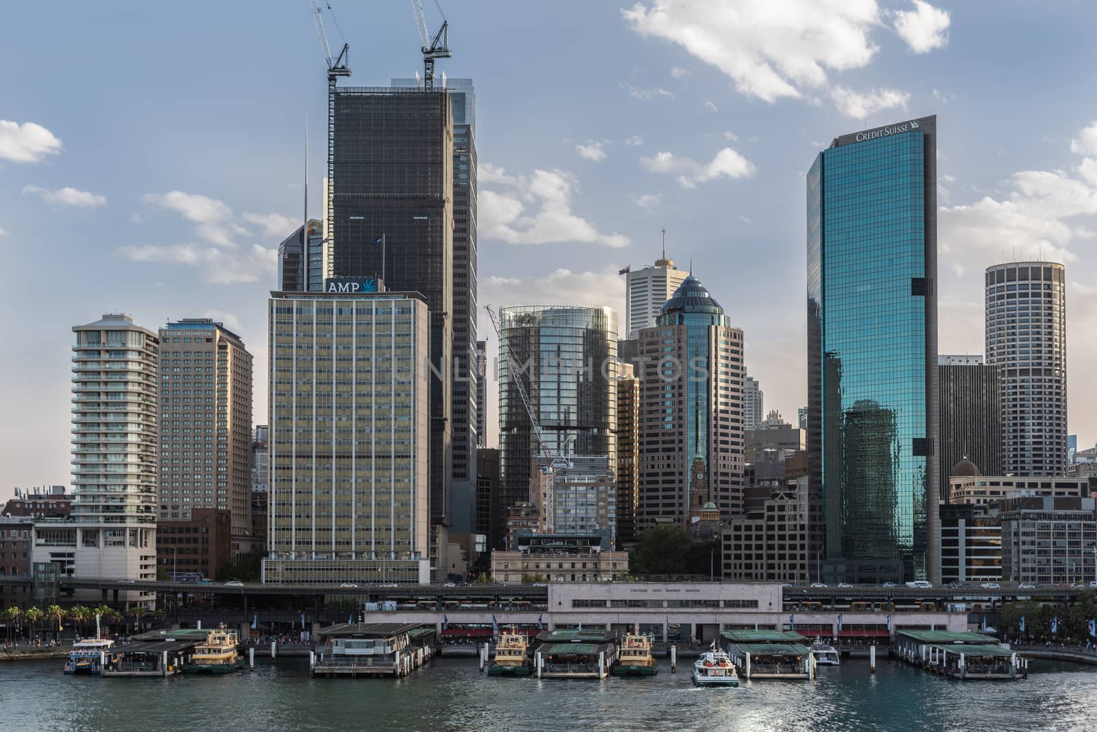 Sydney, Australia - February 12, 2019: Ferry terminal and Circular Quay Railway Station plus skyline in back. Highrises under construction with cranes. Evening shot with light blue sky.