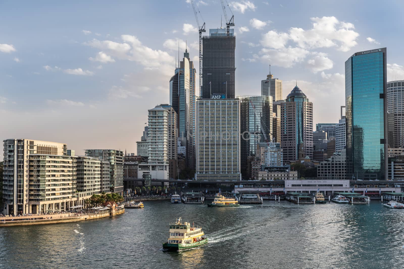 Sydney, Australia - February 12, 2019: Eastern side of Ferry terminal and Circular Quay Railway Station plus skyline in back. Highrises under construction with cranes. Evening shot with light blue sky.
